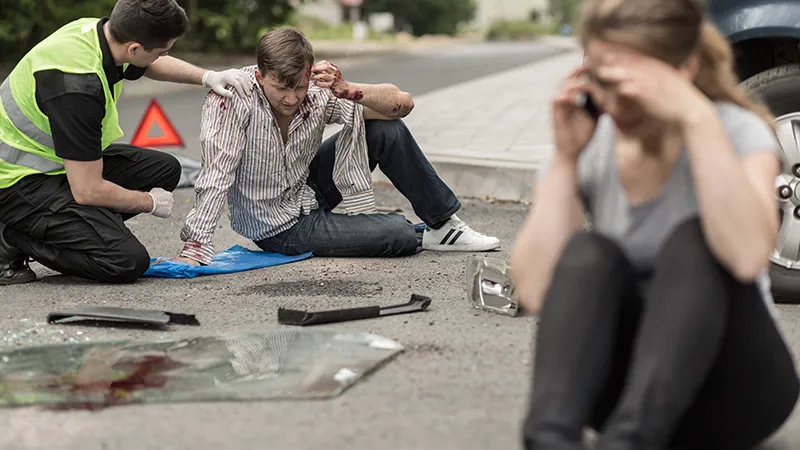 The injured man has blood on his face and shirt, and he appears to be in pain. In the foreground, a woman is sitting on the ground, seemingly distressed and possibly on the phone, with her hand covering her face.